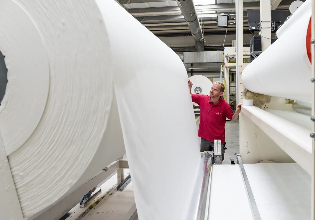 Es ist die Produktion von Vliesstoffen zu sehen. Das Vlies läuft über eine große Rolle und geht über eine Mangel zur nächsten Rolle über. Ein Mitarbeitender kontrolliert die Produktion.